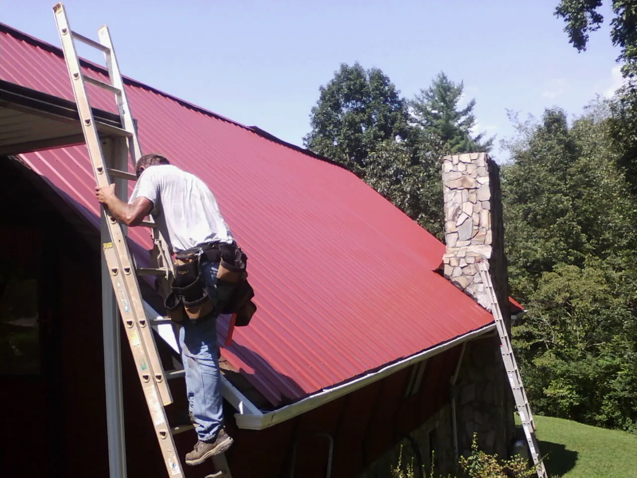 Roofer on a ladder beside a red metal roof and stone chimney — Soffit & Fascia Repair by Roofing Brothers in West Alton